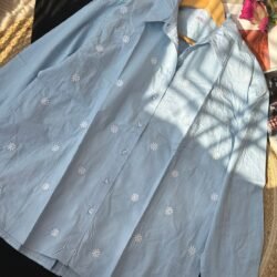 Light blue cotton shirt with white daisy embroidery laid flat with black trousers