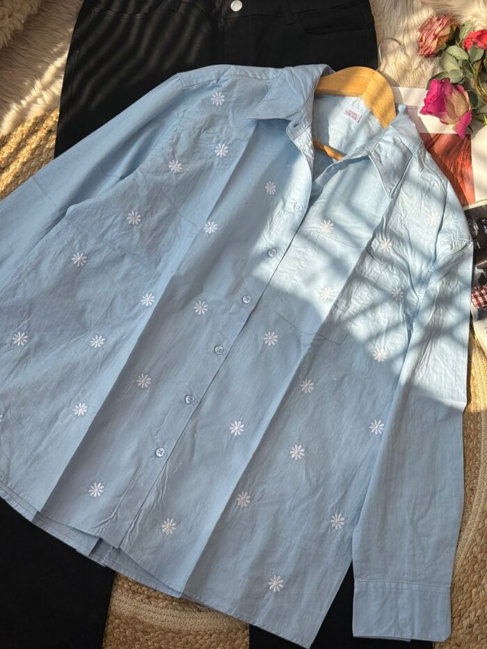 Light blue cotton shirt with white daisy embroidery laid flat with black trousers